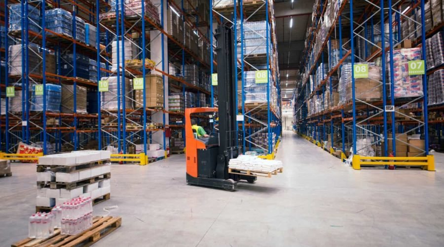 Un magasinier utilise un chariot élévateur pour déplacer des palettes de marchandises entre les rayonnages d'un grand entrepôt organisé.