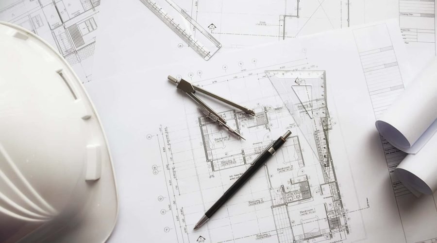 Plans d'architecte avec un crayon, un compas, une règle et un casque de sécurité blanc sur un bureau ; certains plans sont partiellement enroulés.