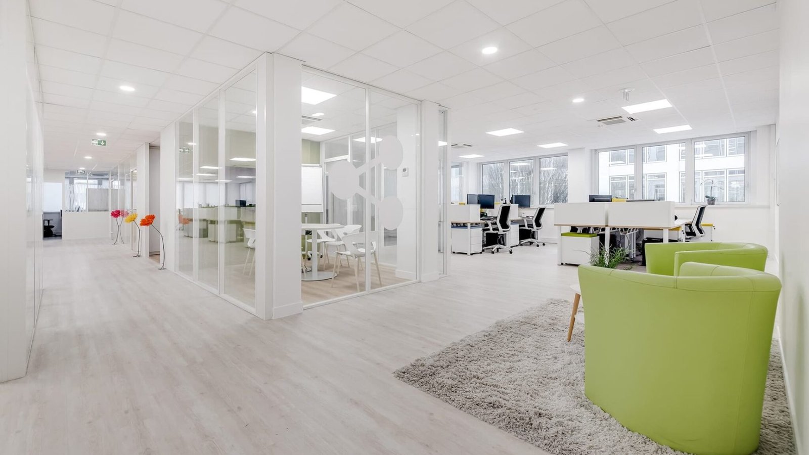 Espace de bureau moderne avec des salles de réunion en verre, des bureaux blancs, des chaises vertes et un tapis gris sur un sol en bois clair, avec de grandes fenêtres apportant de la lumière naturelle.