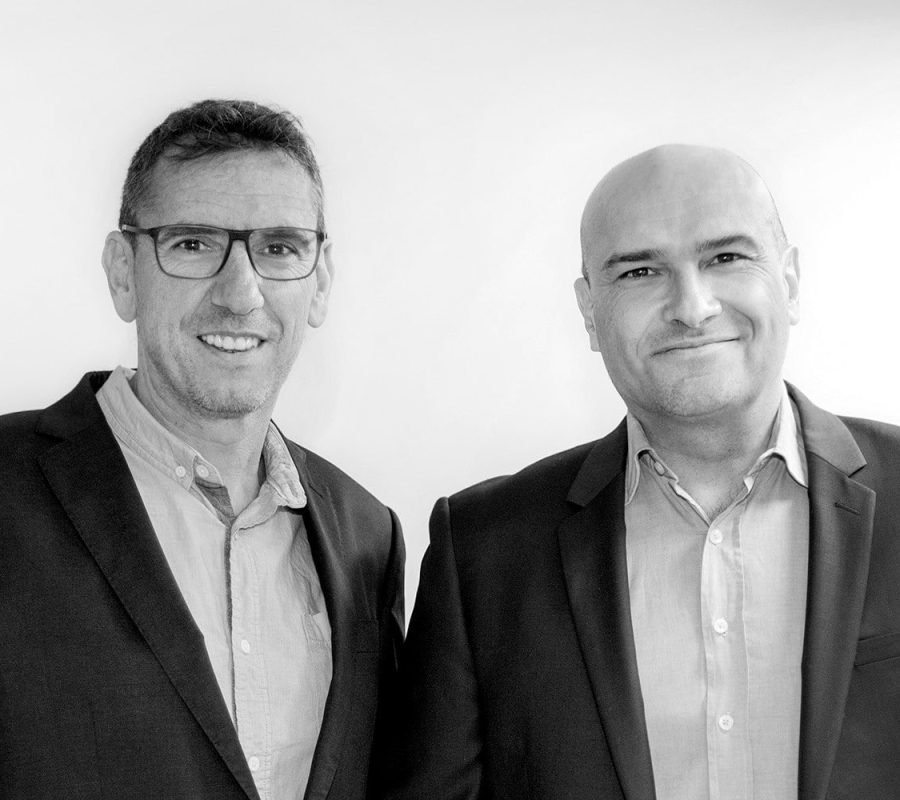 Deux hommes en tenue de travail se tiennent côte à côte sur un fond uni, tous deux face à la caméra et souriants.