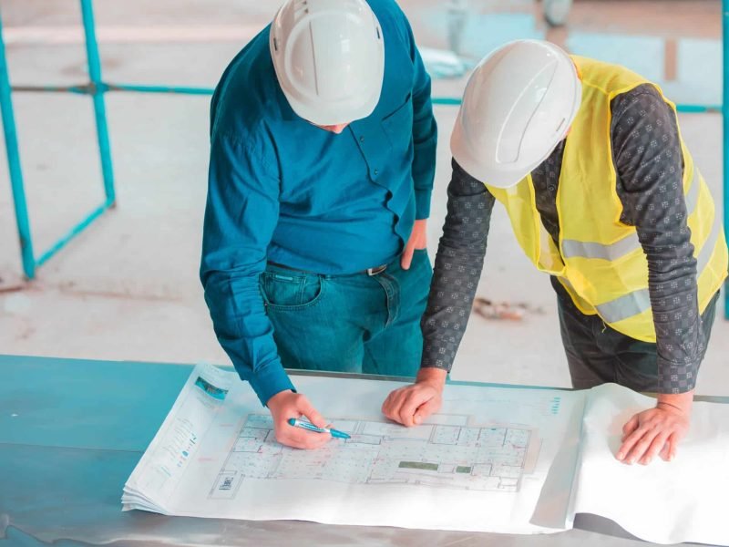 Deux personnes portant des casques de sécurité examinent des plans de construction sur une table sur un chantier de construction ; l'une d'elles pointe les plans avec un stylo.