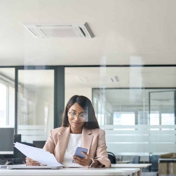 Une femme vêtue d'un blazer léger est assise à un bureau moderne, tenant des documents dans une main et un smartphone dans l'autre, avec des ordinateurs et des murs en verre en arrière-plan.