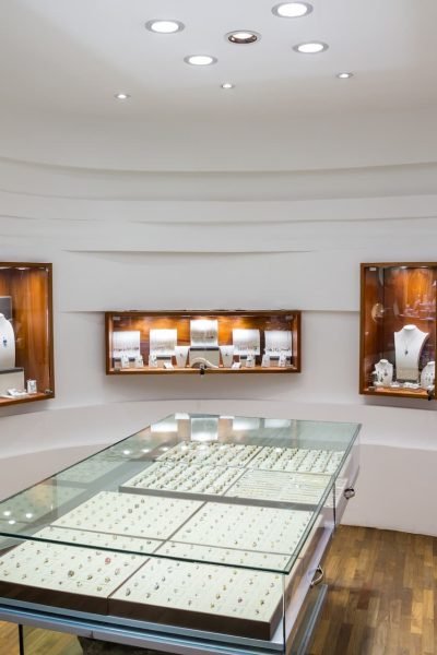 Intérieur d'une bijouterie avec des vitrines en verre présentant des bagues et des vitrines murales présentant des colliers et d'autres bijoux, sur un fond blanc et moderne.