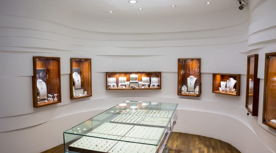 Intérieur d'une bijouterie avec des vitrines en verre présentant des bagues et des vitrines murales présentant des colliers et d'autres bijoux, sur un fond blanc et moderne.