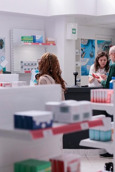 Plusieurs personnes interagissent au comptoir d'une pharmacie, avec des étagères de médicaments et de produits de santé au premier plan.