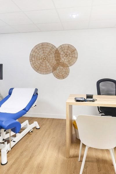 Une salle d'examen médical moderne avec une table d'examen bleue, un appareil à ultrasons, un bureau avec des chaises, un ordinateur et des œuvres d'art murales décoratives.