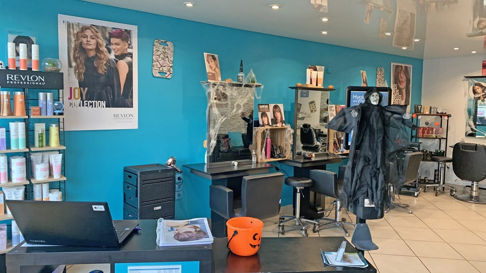 Un salon de coiffure avec des murs bleus, des présentoirs de produits capillaires, des décorations d'Halloween, notamment une sorcière suspendue et un seau à citrouille, et des postes de coiffure avec des chaises et des miroirs.