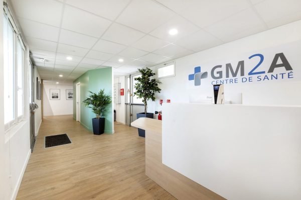 Réception d'un centre médical avec un bureau blanc, des plantes d'intérieur, un sol en bois clair et un panneau indiquant "GM2A Centre de Santé" sur le mur.