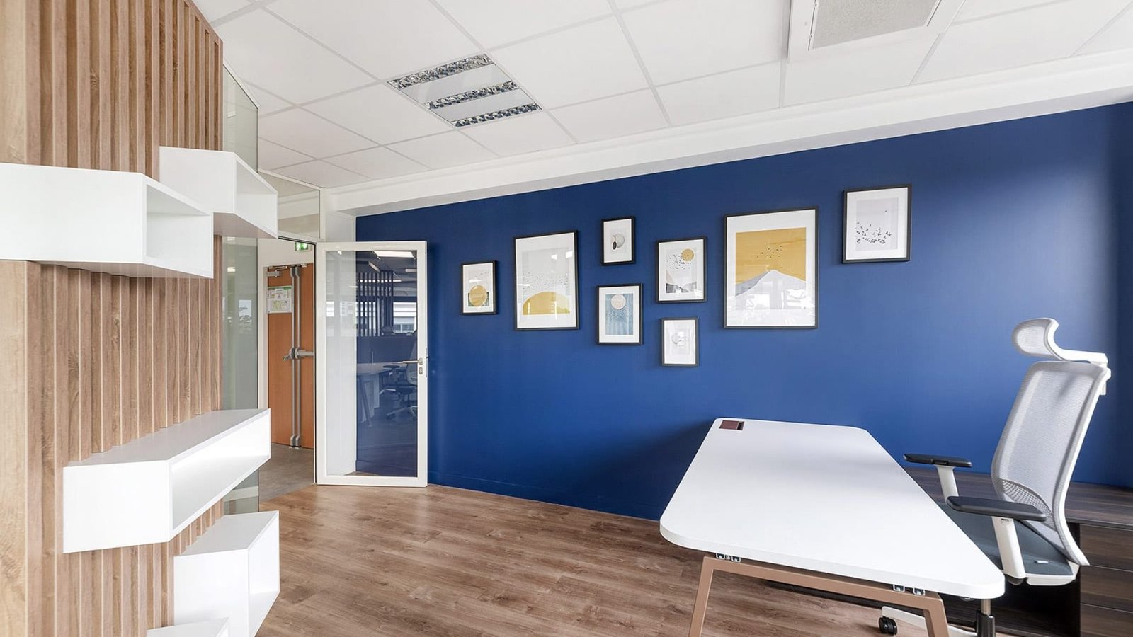 Bureau moderne avec un mur d'accent bleu, des œuvres d'art encadrées, un bureau blanc, une chaise ergonomique, un sol en bois et des étagères. Une porte vitrée mène à une autre pièce.