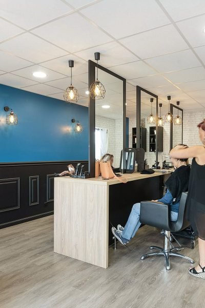 Une coiffeuse coiffe une cliente dans un salon moderne aux murs bleus et blancs, au sol en bois et aux étagères présentant des produits capillaires.