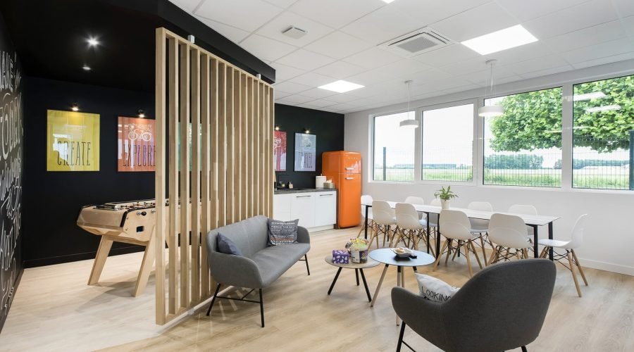 Salle de pause moderne avec un baby-foot, un coin salon, une table à manger, un réfrigérateur orange et de grandes fenêtres laissant entrer la lumière naturelle.