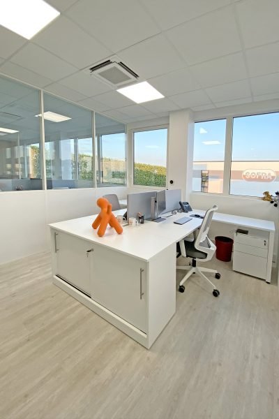 Bureau moderne avec un bureau blanc, des écrans d'ordinateur, une chaise de bureau et une sculpture orange en forme de chien près de grandes fenêtres.