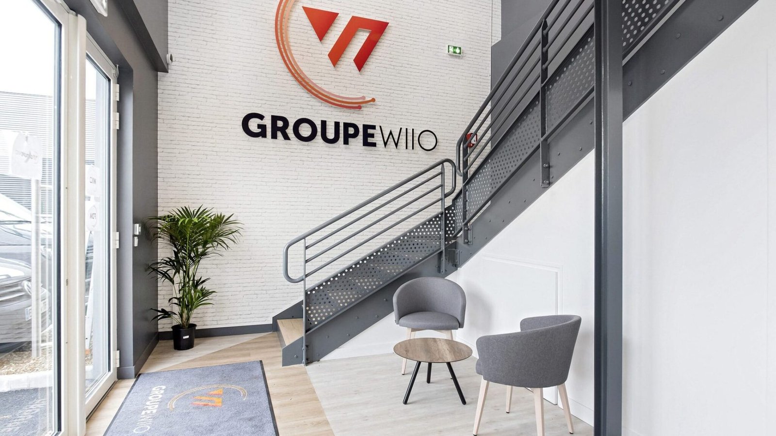 Entrée de bureau moderne avec deux chaises grises, une petite table ronde, une plante en pot et un escalier. Logo "GROUPE WIO" affiché sur un mur de briques blanches.